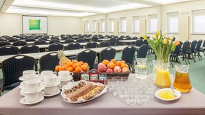 Un salón de conferencias con filas de sillas, mesas con alimentos para el desayuno y una pantalla de proyector al fondo.