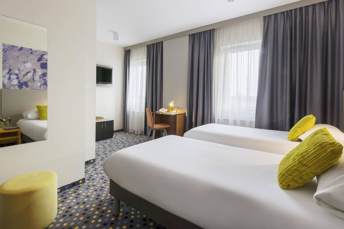 Een hotelkamer met twee bedden, een bureau, een stoel, een televisie en een groot raam met gordijnen.