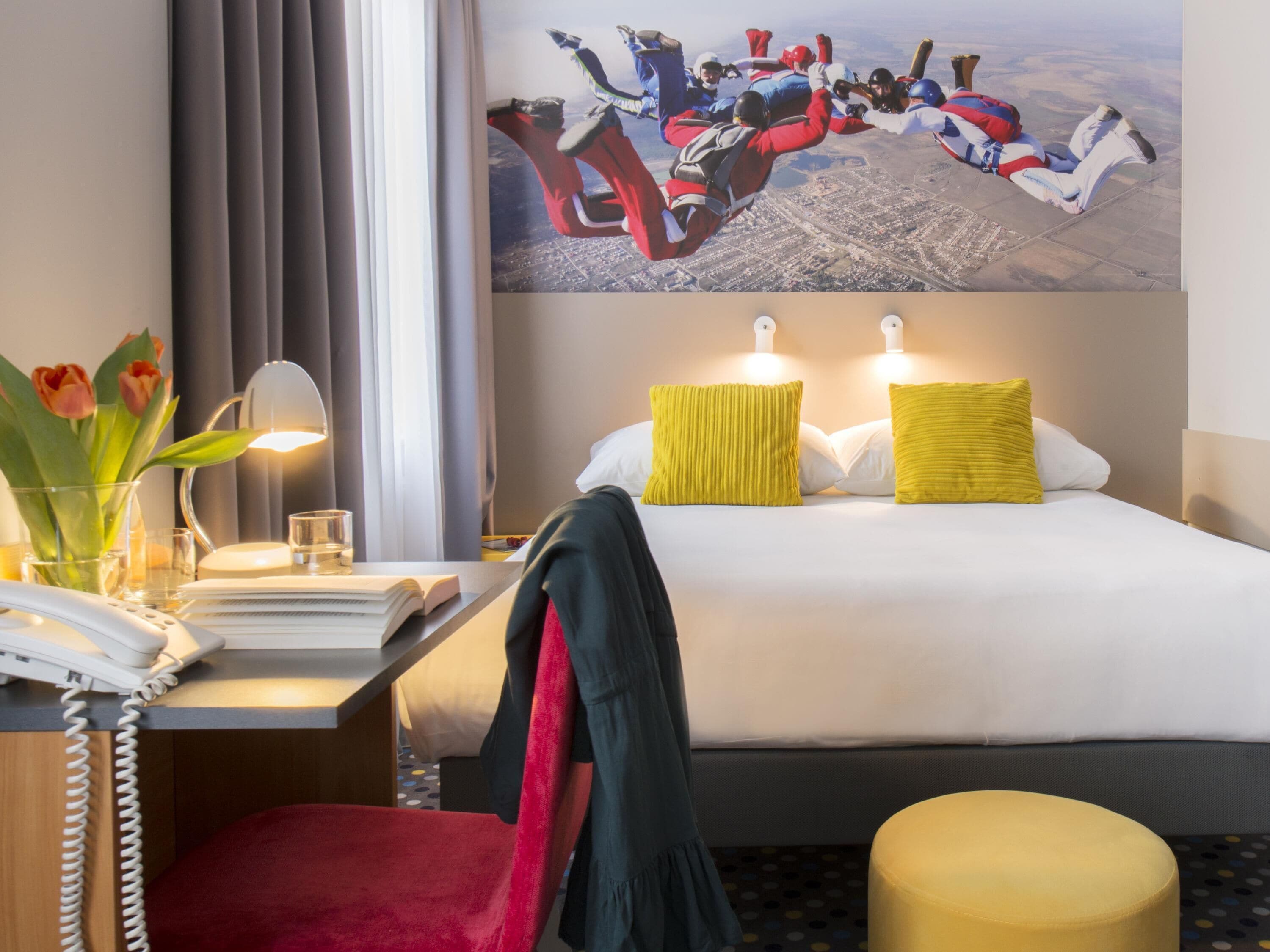 A hotel room with a bed, bedside table, lamp, and a painting of skydivers.