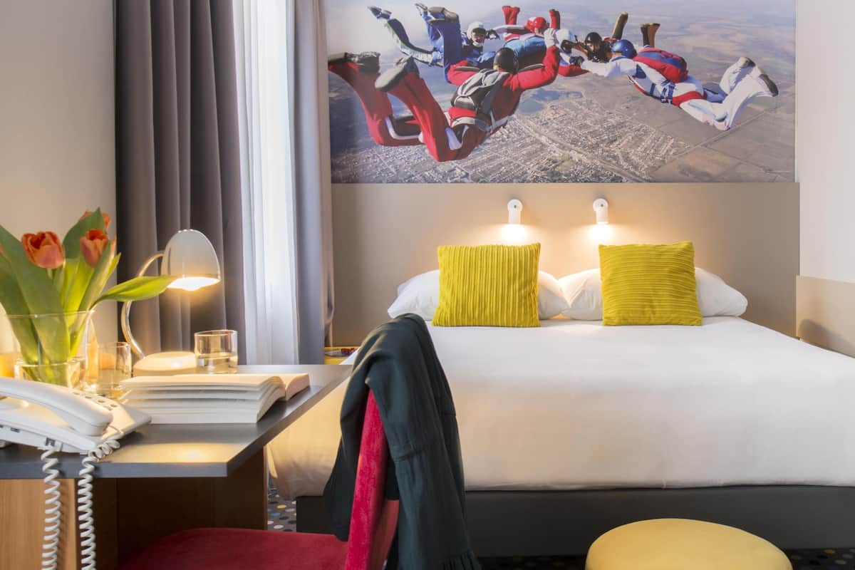 Een hotelkamer met een bed, nachtkastje, lamp en een schilderij van skydivers.