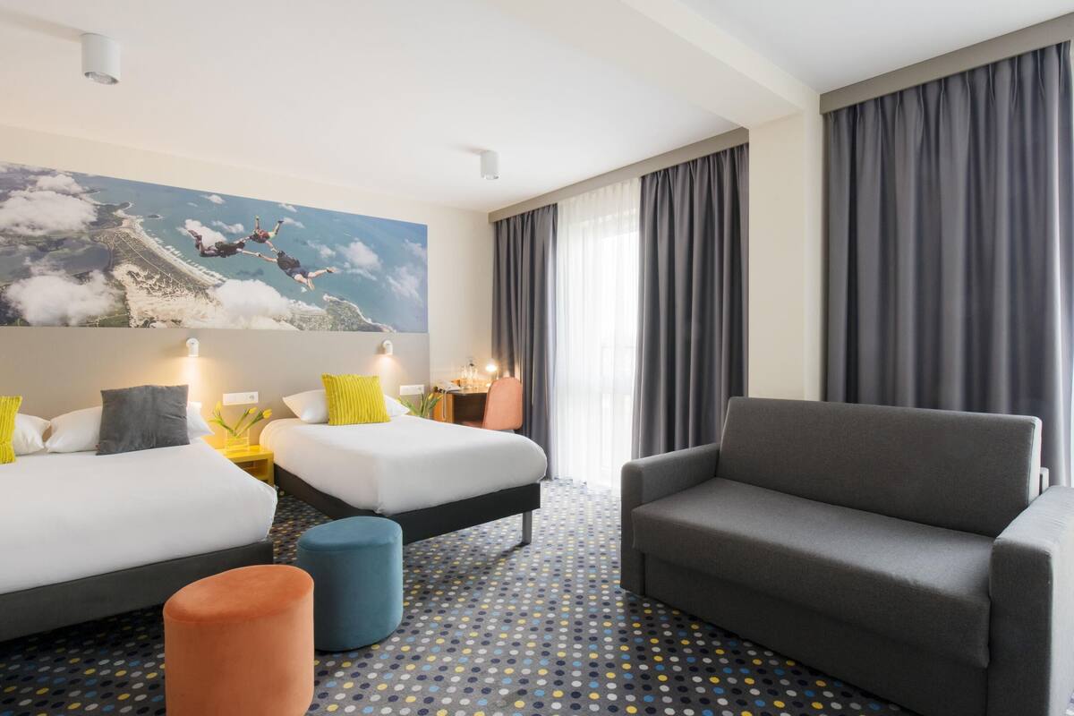 Habitación de hotel con dos camas, un sofá, un pequeño otoman, un escritorio y un ventanal con cortinas.
