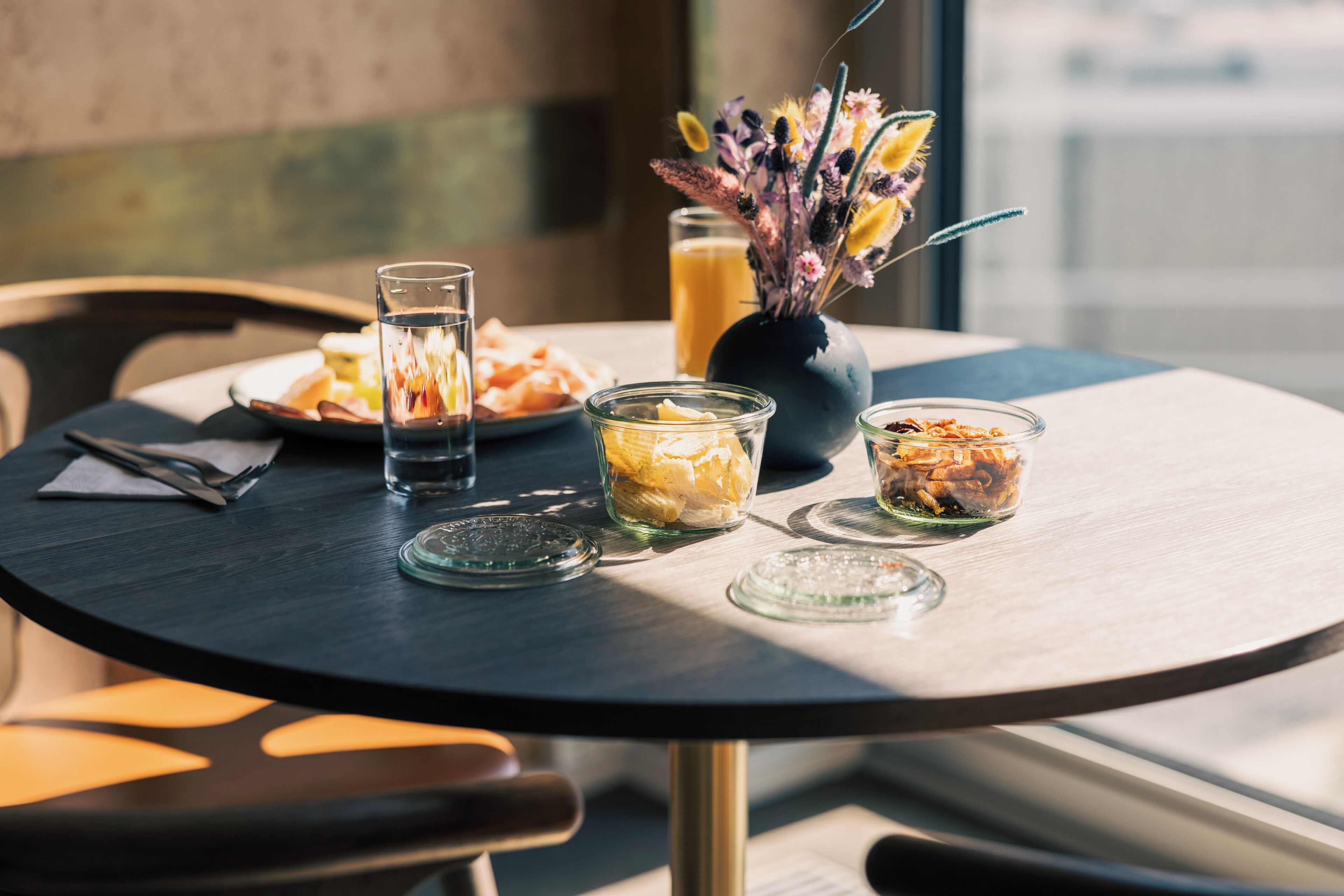 A round table with a glass of water, a glass of orange juice, and two small glass jars with food items. There is a vase with flowers in the background.