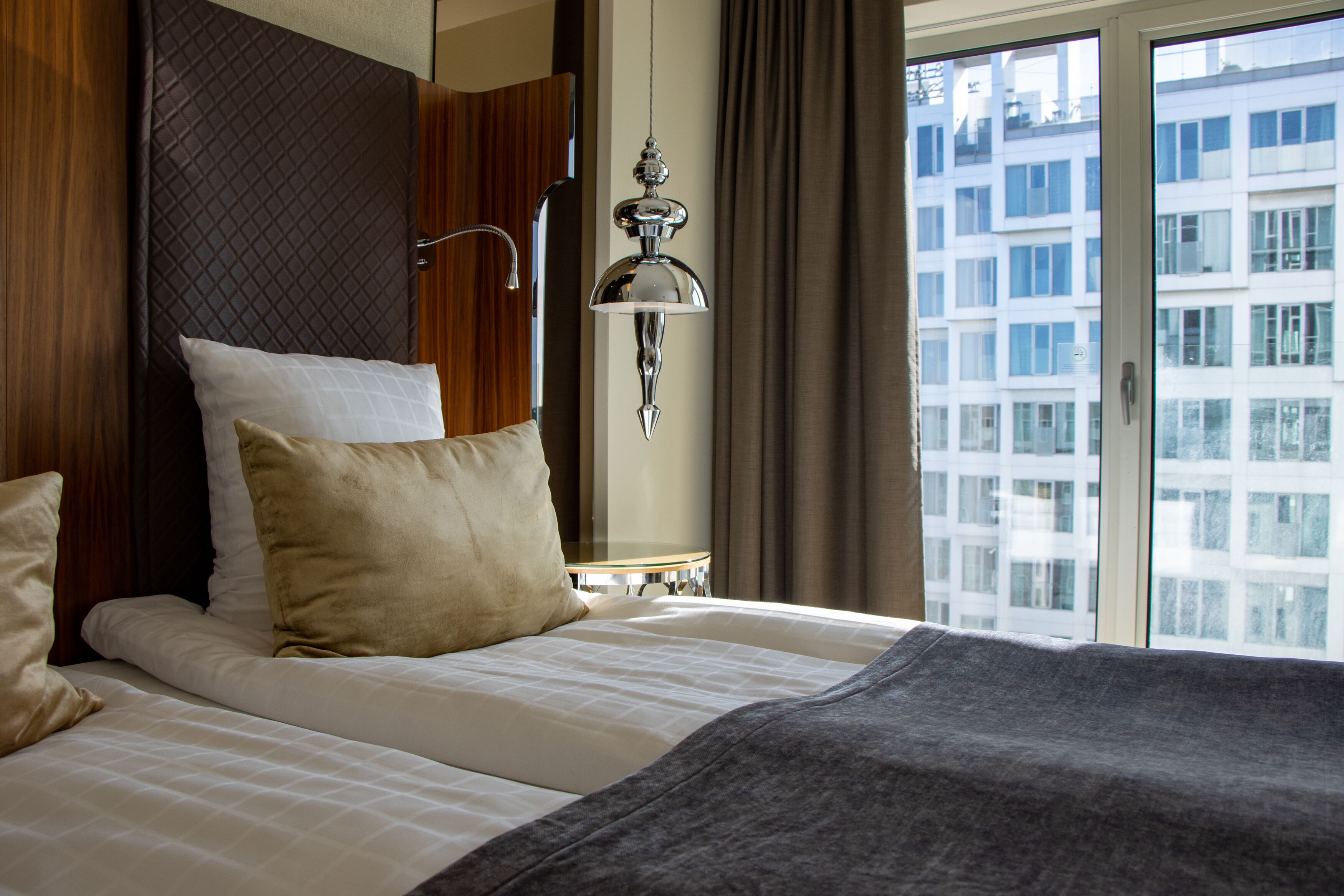 A hotel room with a bed, a nightstand, a lamp, and a view of buildings through a window.