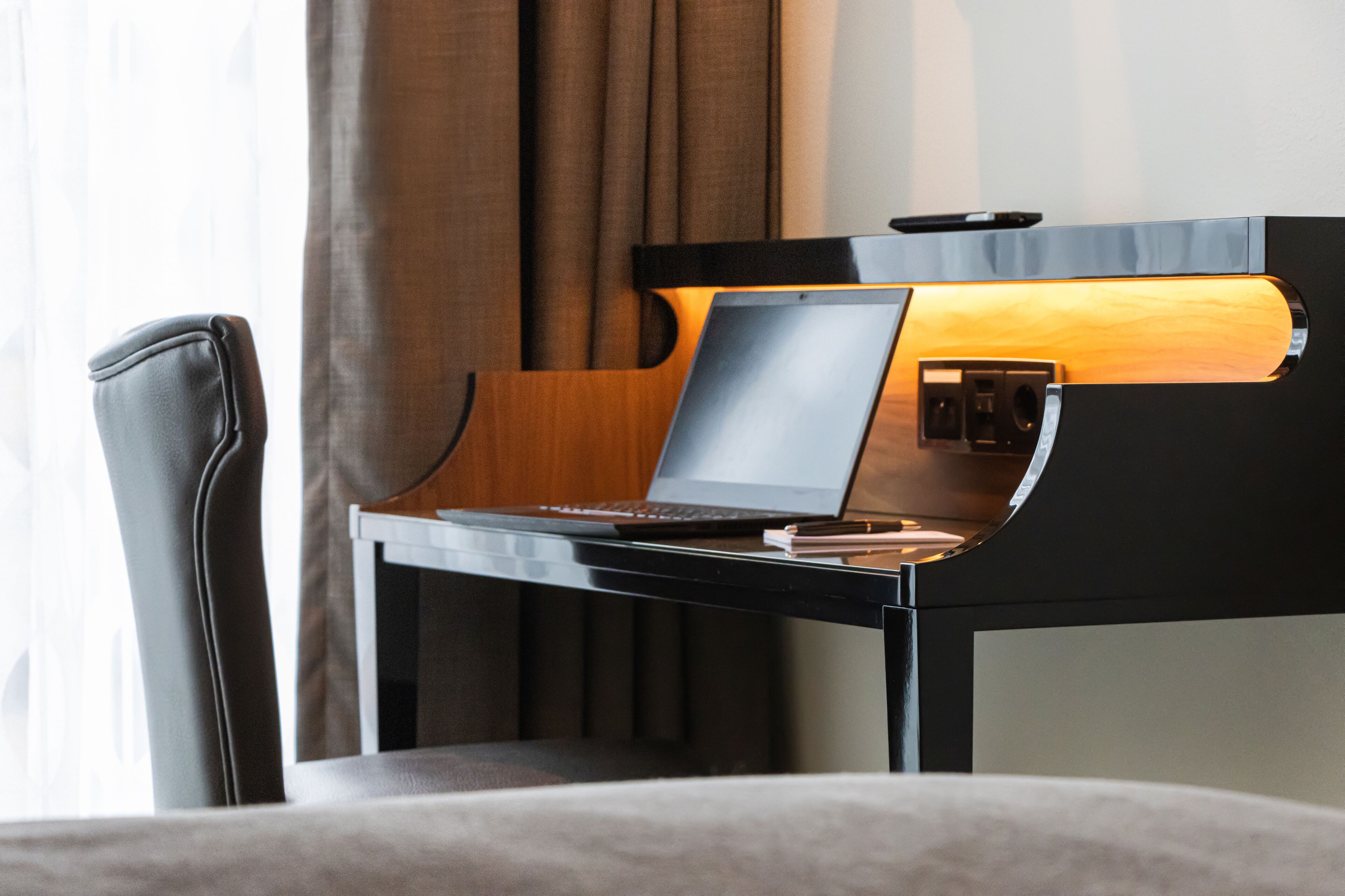 Moderni hotellihuone, jossa on tyylikäs musta työpöytä, kannettava tietokone ja lamppu.
