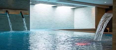 Una piscina interior moderna con dos cascadas, suelo de baldosas y techo alto con iluminación empotrada.