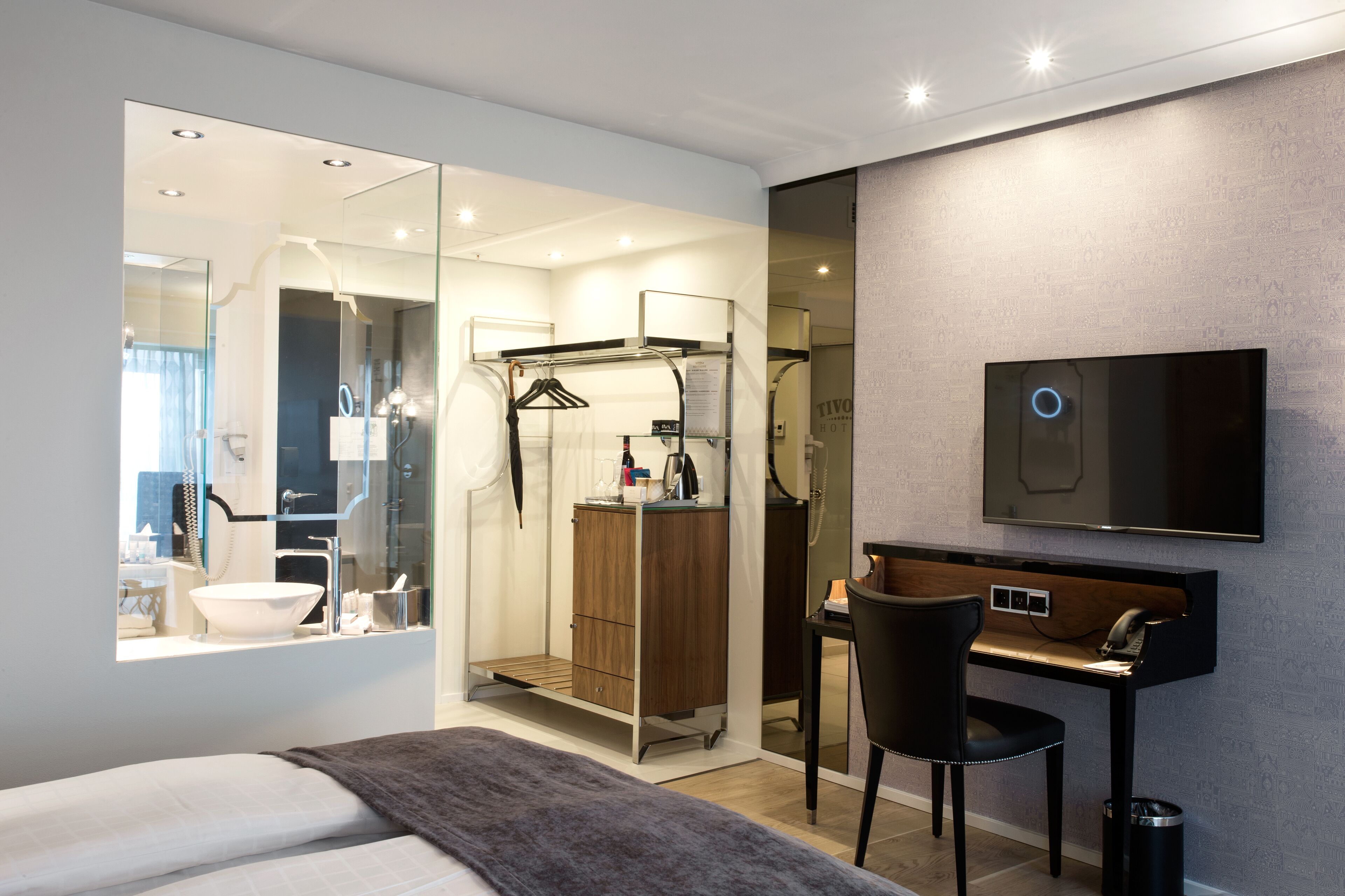Habitación de hotel moderna con una cama grande, un escritorio con silla, un televisor montado en la pared y un espejo que refleja el baño.