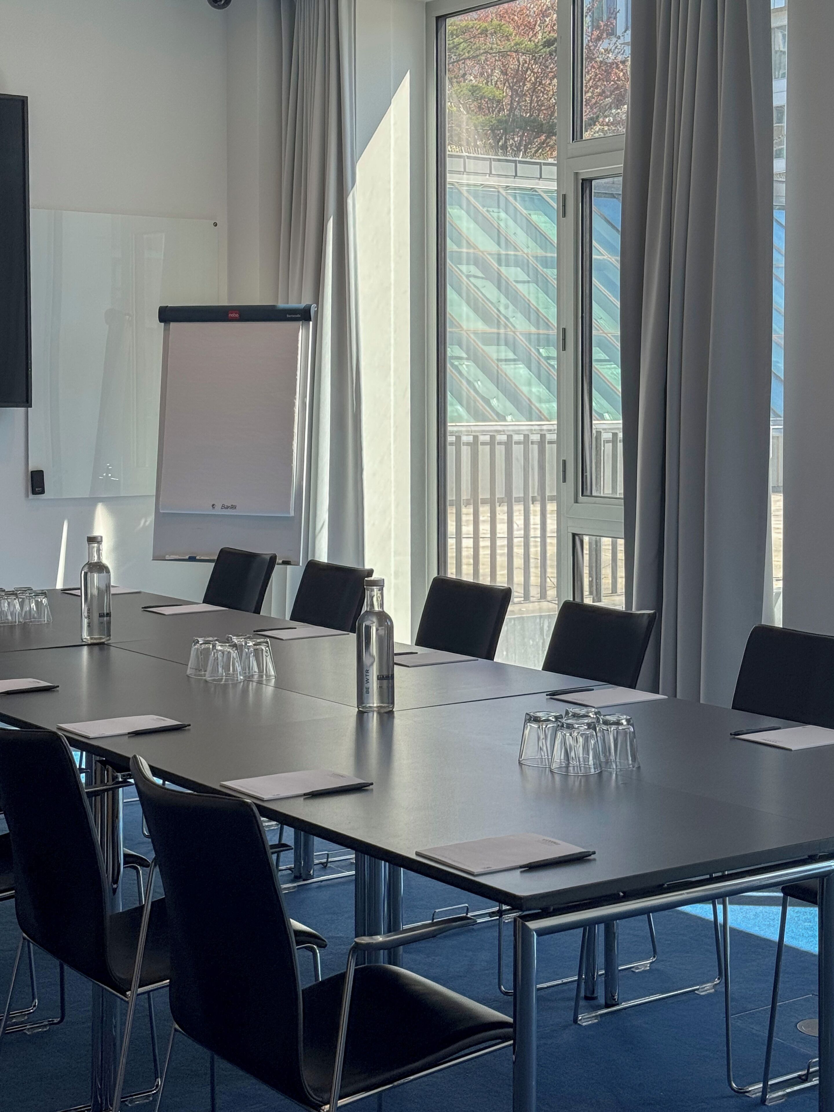 A conference room with a long table, chairs, a projector screen, and a view of a building through large windows.