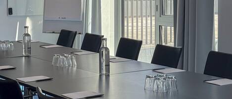 A conference room with a long table, chairs, a projector screen, and a view of a building through large windows.