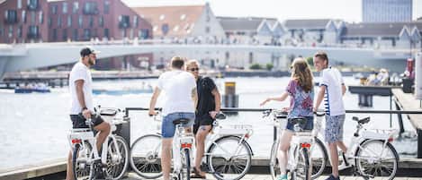Un grupo de personas en un muelle de madera junto a un río, con bicicletas y un puente al fondo.