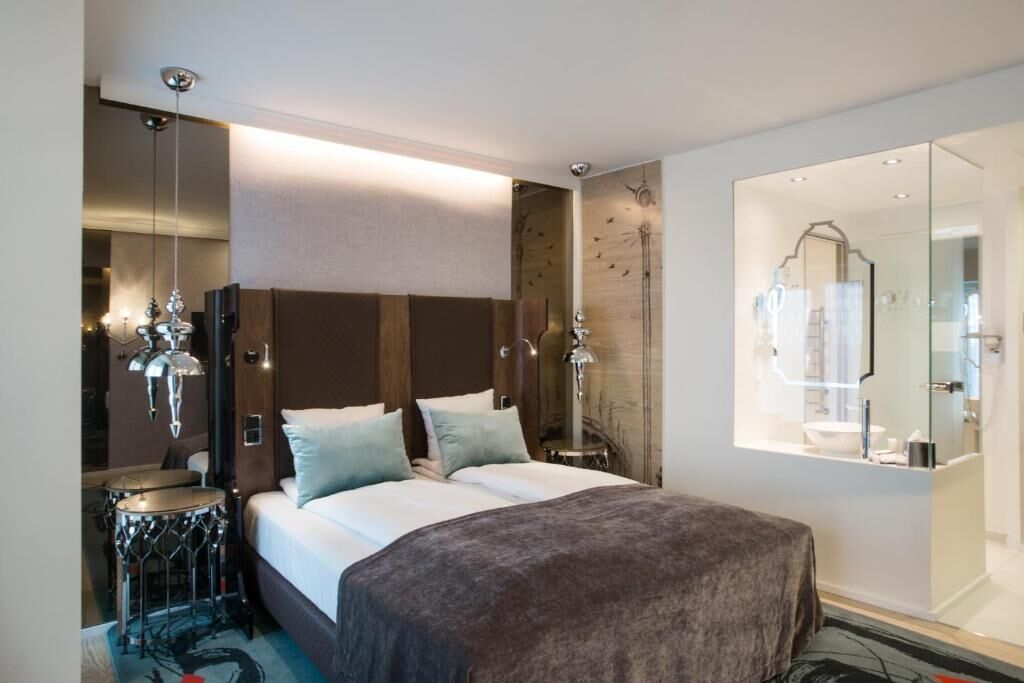 A modern hotel room with a large bed, a drum set, a mirror, and a well-lit bathroom.