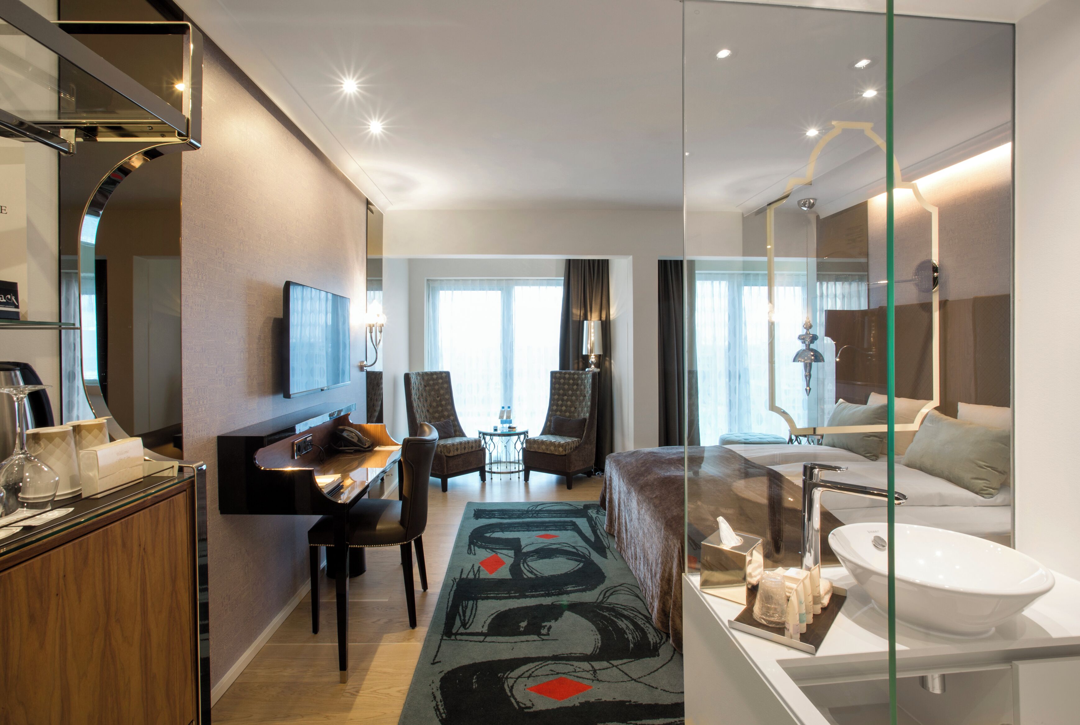 A modern hotel room with a glass partition, a flat-screen TV, a sofa, a dining area, and a bathroom with a sink and mirror.