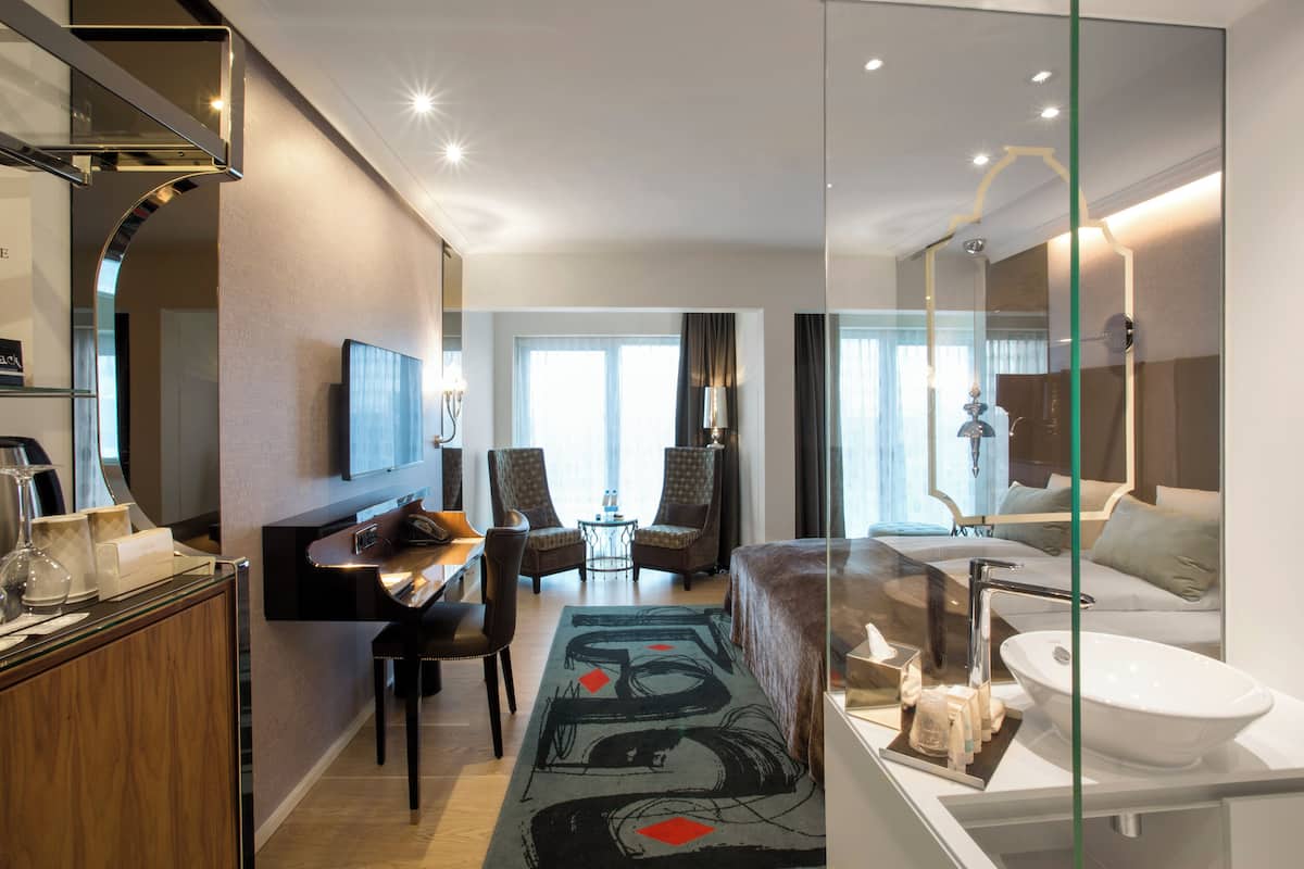 Quarto de hotel moderno com divisória de vidro, TV de tela plana, sofá, área de refeições e banheiro com pia e espelho.
