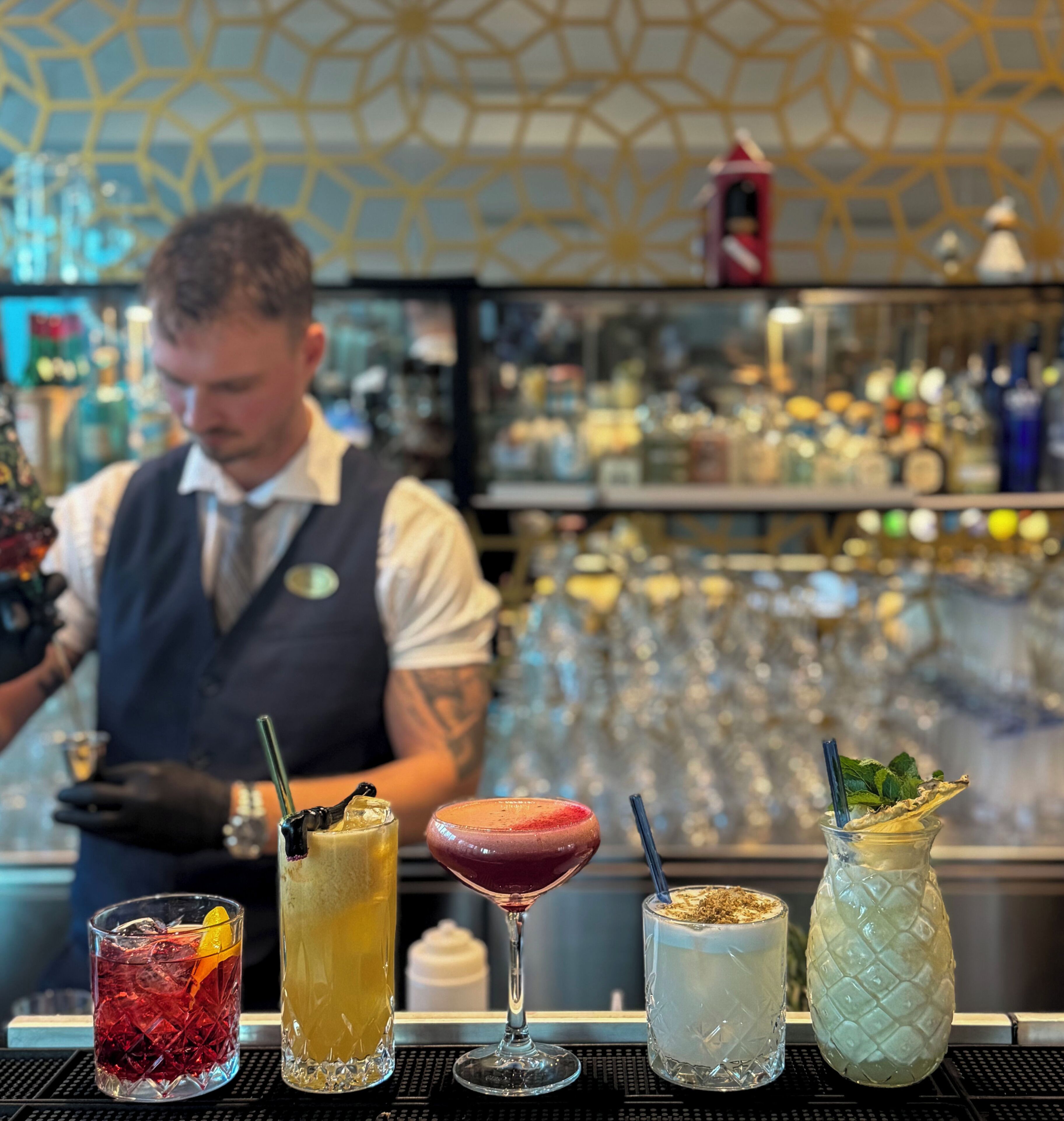 A bar area with a bartender, a variety of drinks, and a patterned wall in the background.