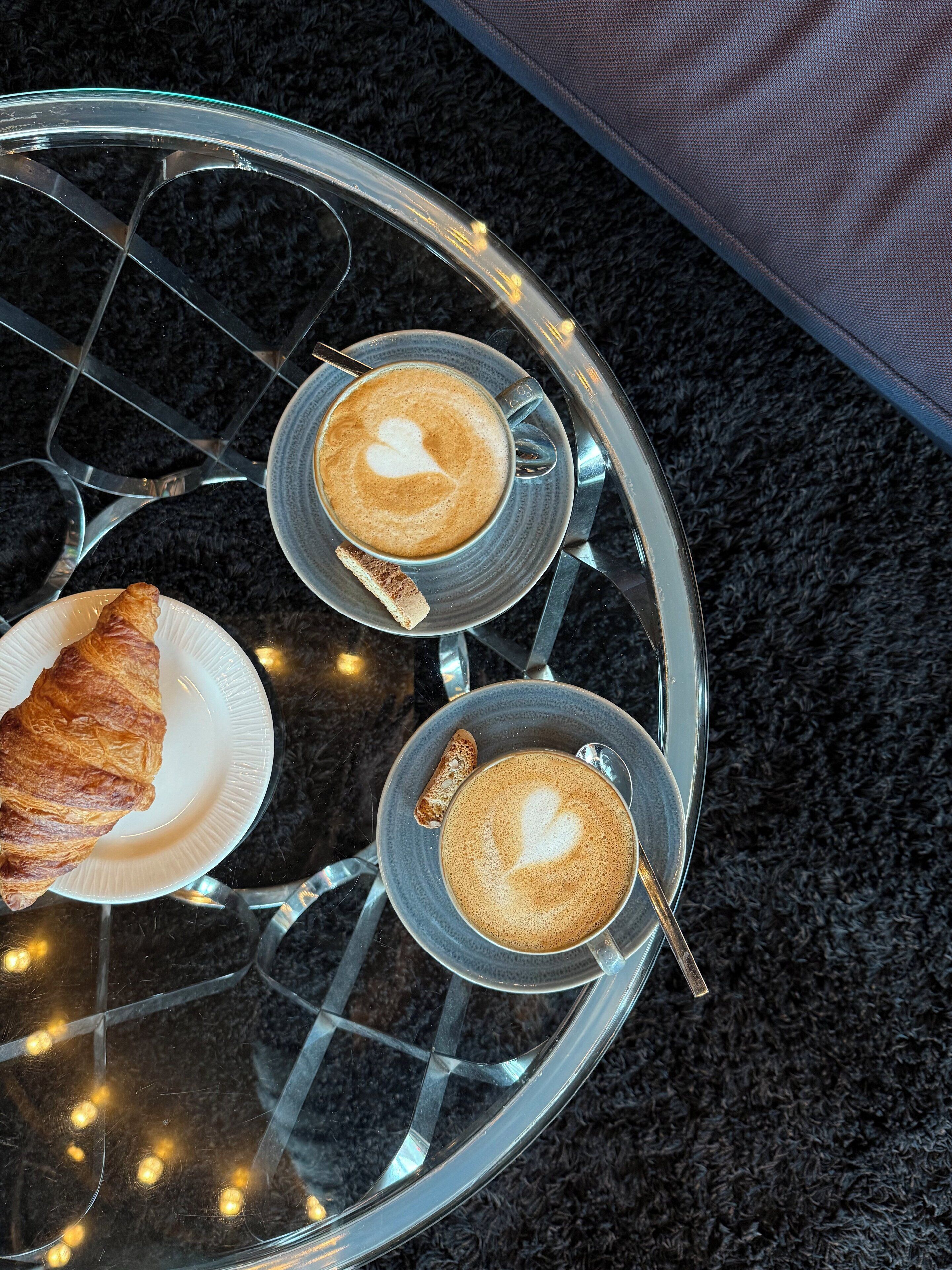 Ein Glastisch mit zwei Kaffeetassen und einem Croissant auf einem dunklen Teppichboden.
