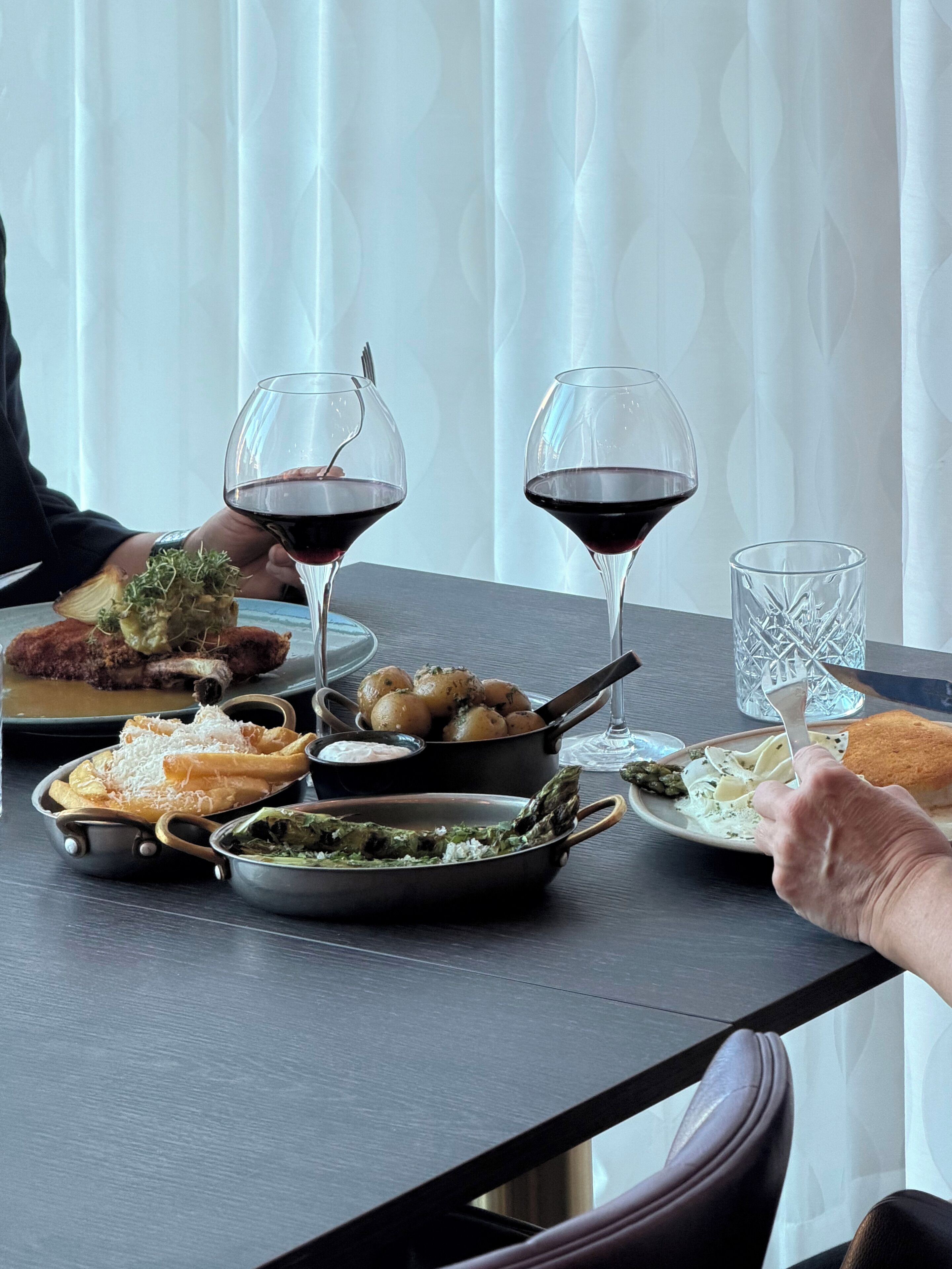 Een eettafel met verschillende gerechten, twee wijnglazen en een persoon die naar een glas reikt.