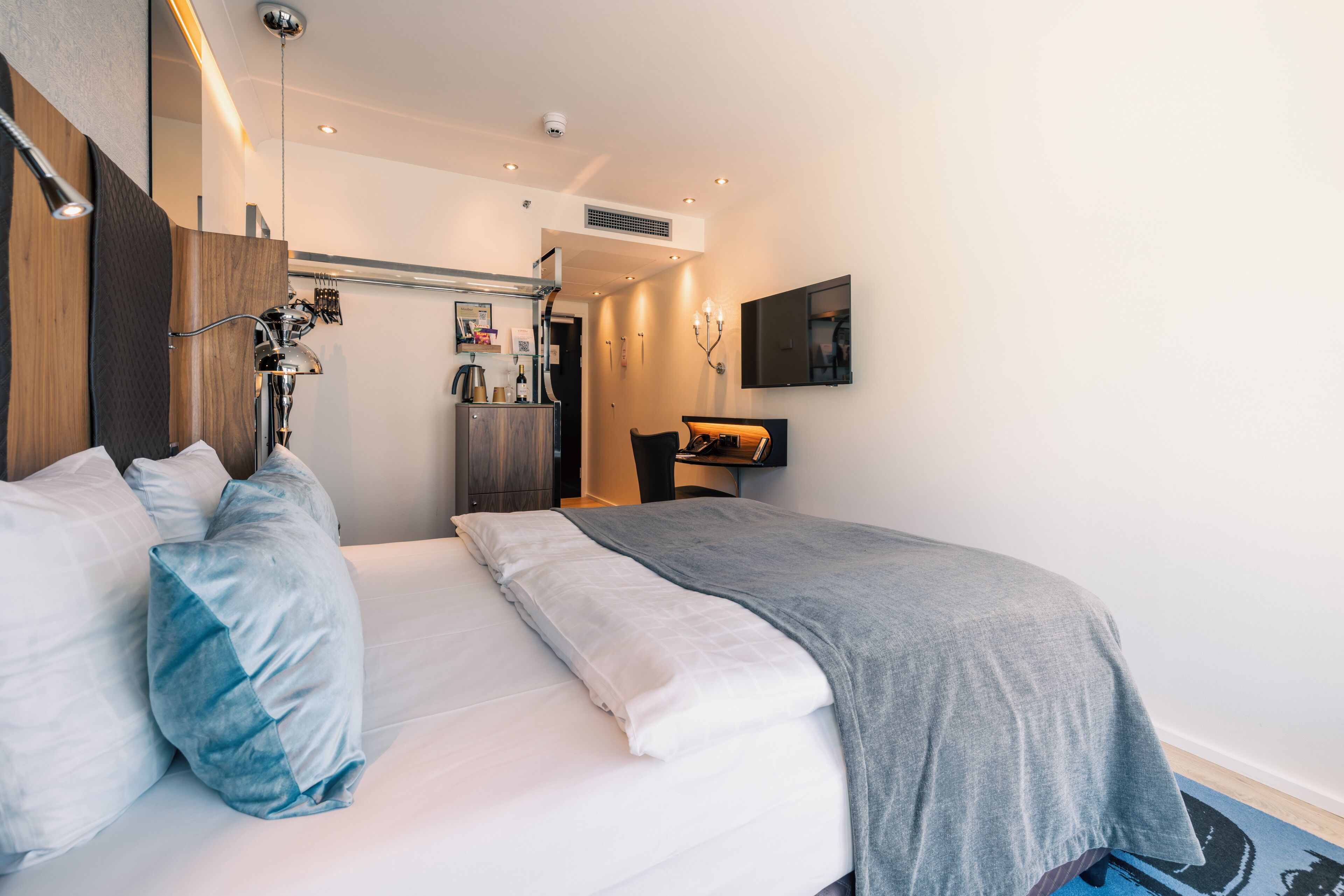 A modern hotel room with a neatly made bed, a desk with a chair, a mounted TV, and a wall-mounted heater.