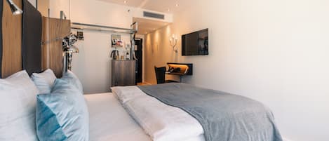 A modern hotel room with a neatly made bed, a desk with a chair, a mounted TV, and a wall-mounted heater.