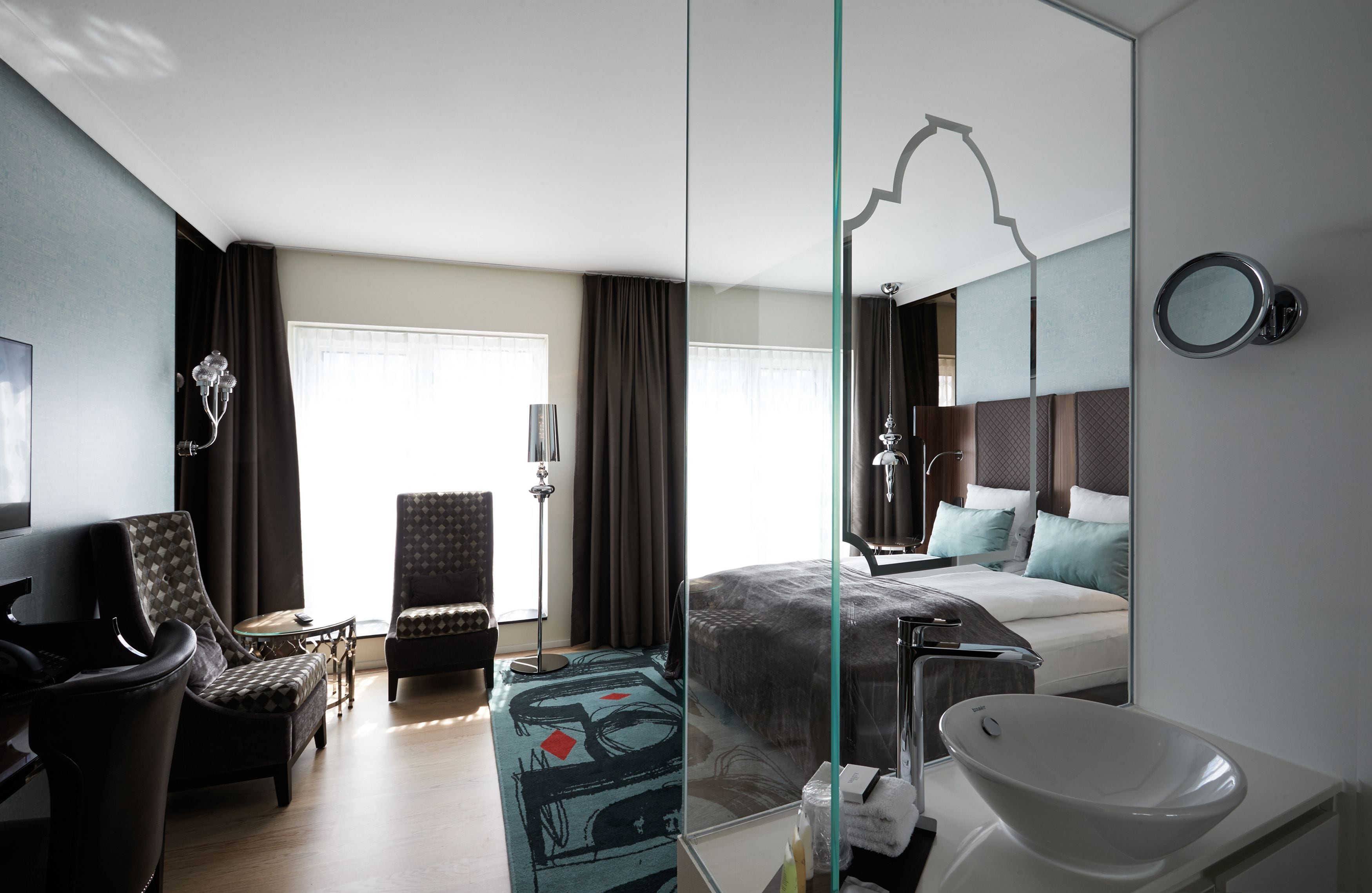 A modern hotel room with a large bed, a sitting area with a chair and a table, a large mirror, and a bathroom with a sink and a large mirror.