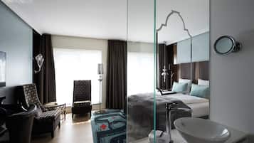 Una moderna camera d'albergo con un letto grande, un'area salotto con una poltrona e un tavolo, uno specchio grande e un bagno con un lavandino e uno specchio ampio.