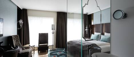 Una habitación de hotel moderna con una cama grande, una zona de estar con una silla y una mesa, un espejo amplio y un baño con lavamanos y un espejo grande.