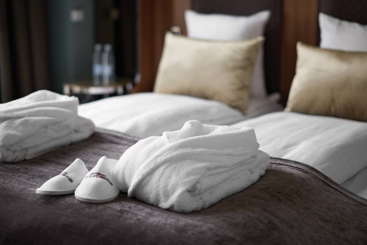A neatly made bed with white linens, a pair of white slippers, and a folded towel on a dark-colored bedspread.