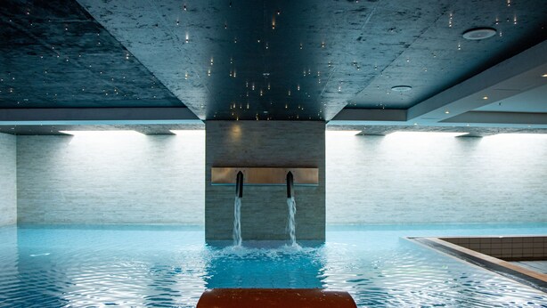 An indoor swimming pool with a modern design, featuring a concrete ceiling and walls, and a central water feature.