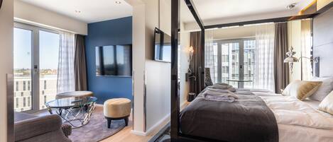 A modern hotel room with a large bed, a TV mounted on the wall, a small round table, and a view of the city through large windows.