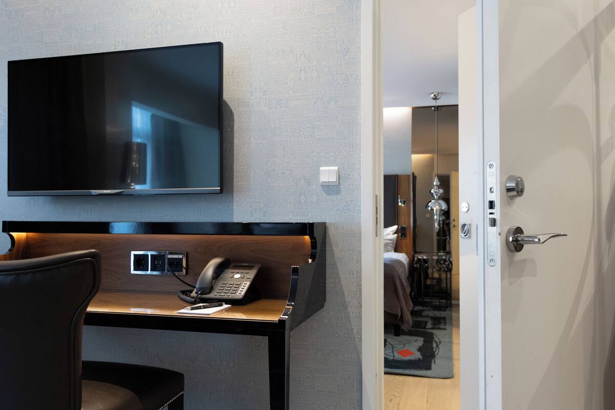 A hotel room with a flat-screen TV mounted on the wall, a desk with a telephone, and a door leading to another room.