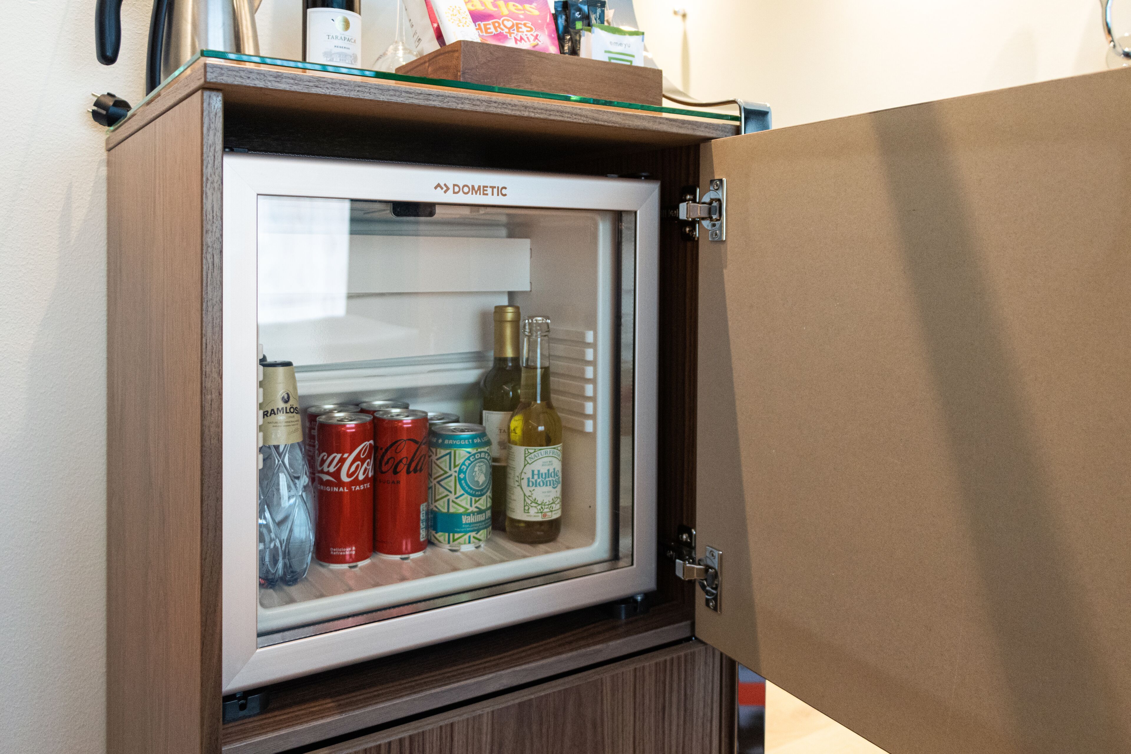 Un refrigerador pequeño con puerta de vidrio, que contiene una botella de vino, una lata de Coca-Cola y una botella de agua.