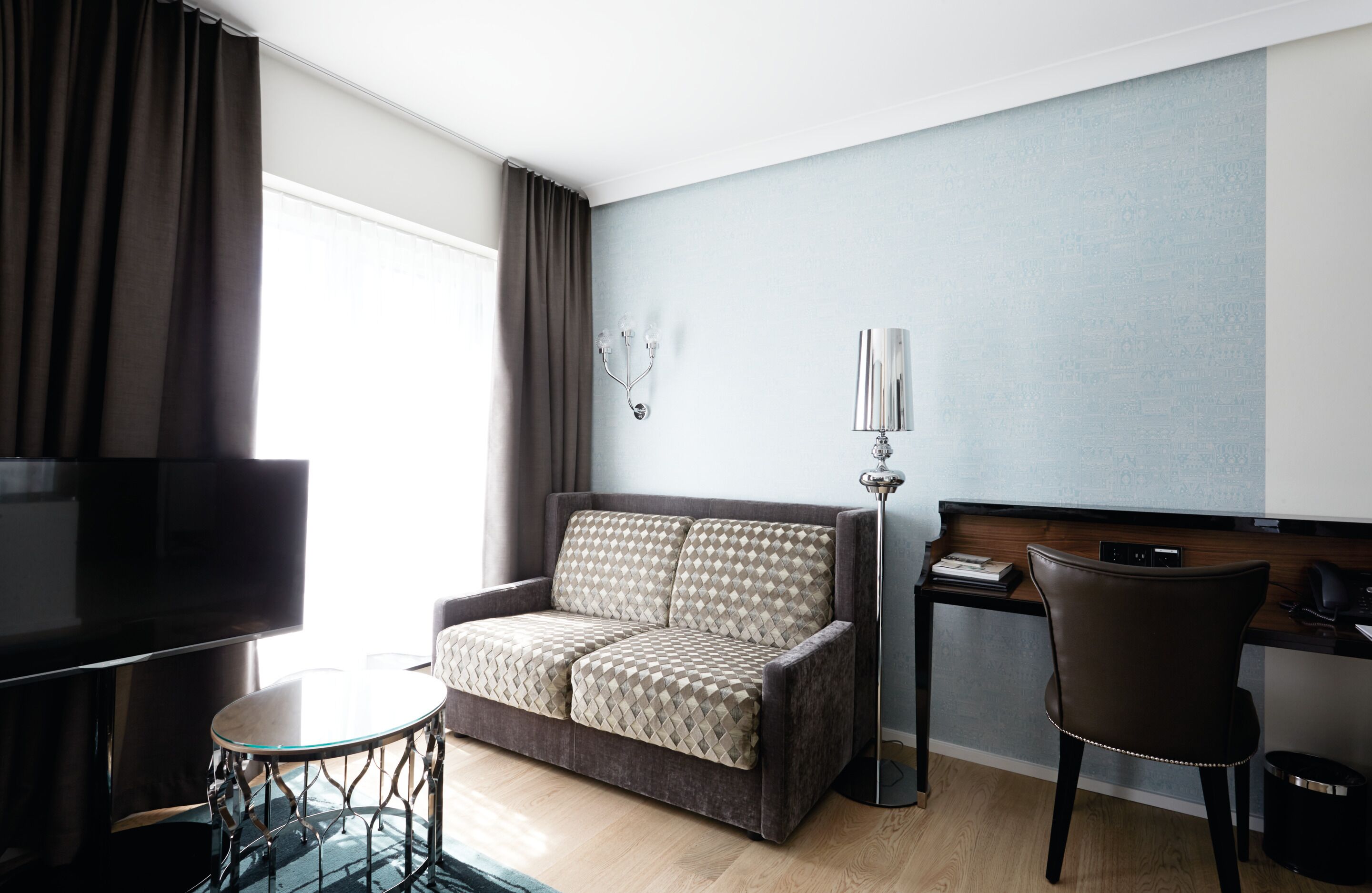 Una habitación de hotel moderna con sofá, una pequeña mesa redonda, televisión, escritorio con silla y lámpara.