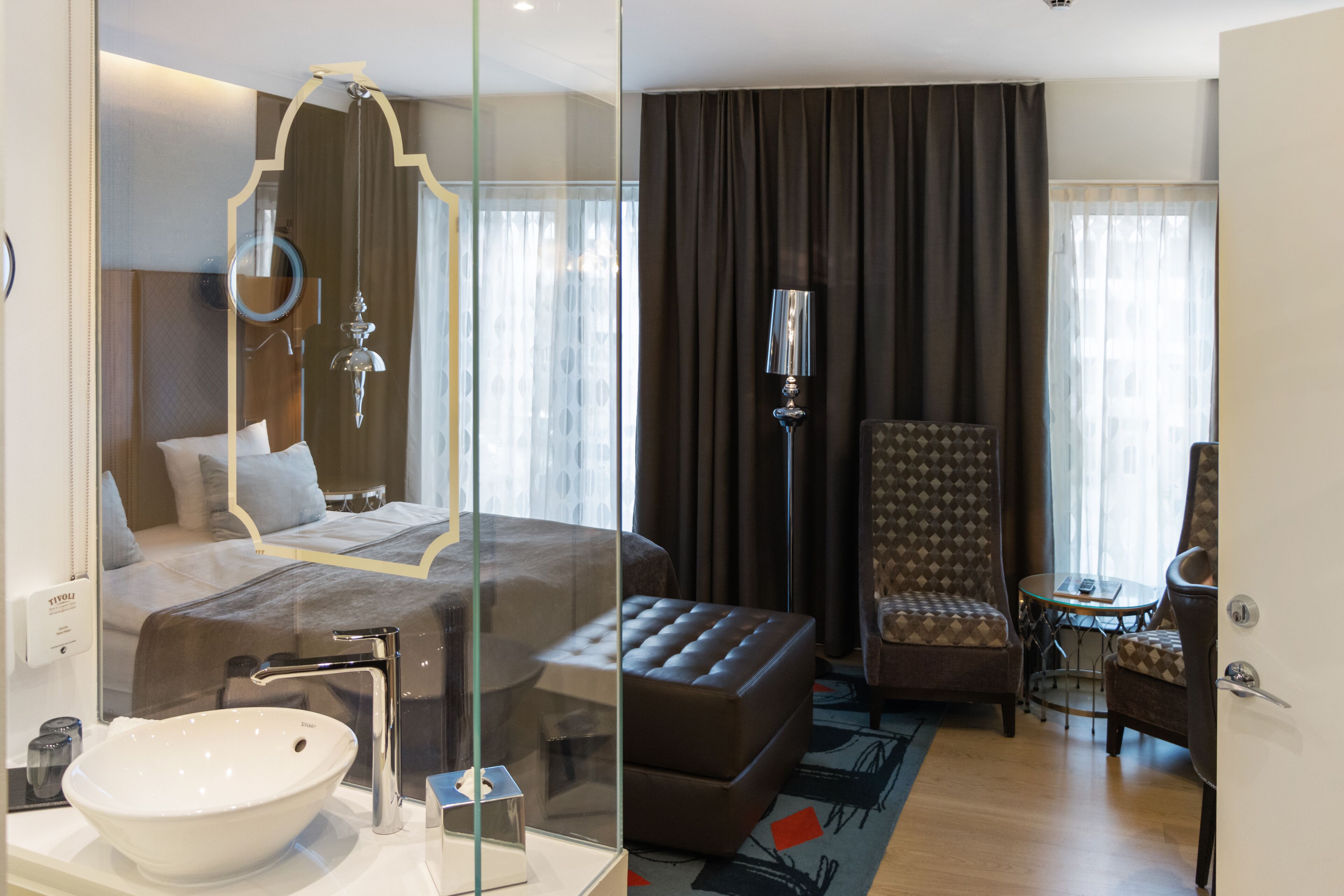 Een moderne hotelkamer met een groot bed, een badkamer met glazen wanden, een zithoek met een tafel en uitzicht op de stad.