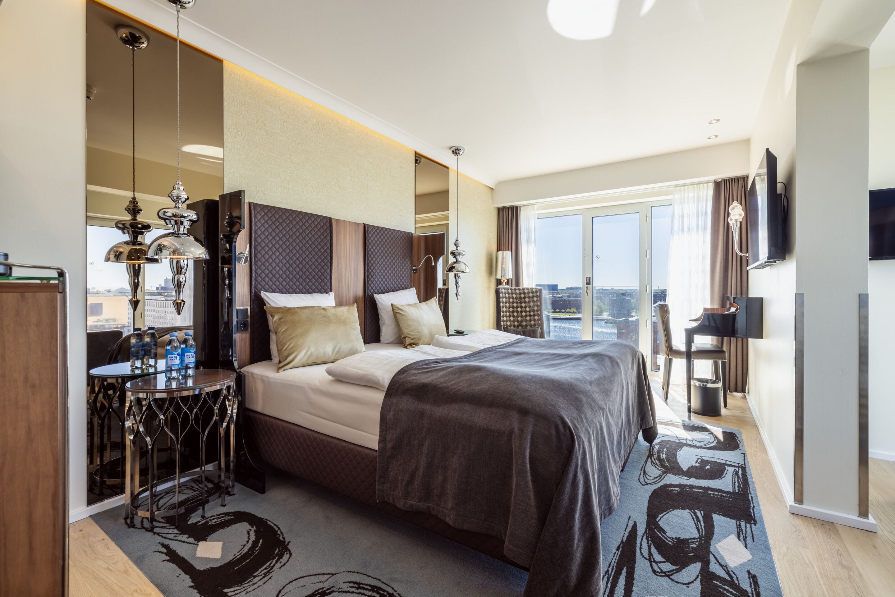 A hotel room with a large bed, a desk, a chair, a TV, and a view of the city.