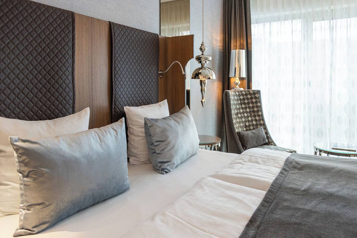 A hotel room with a large bed, multiple pillows, a headboard with a quilted pattern, a wall-mounted lamp, and a chair.
