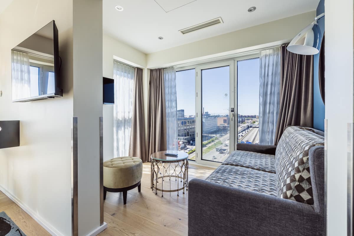 A modern living room with a sofa, a round table, a small ottoman, and a flat-screen TV mounted on the wall.