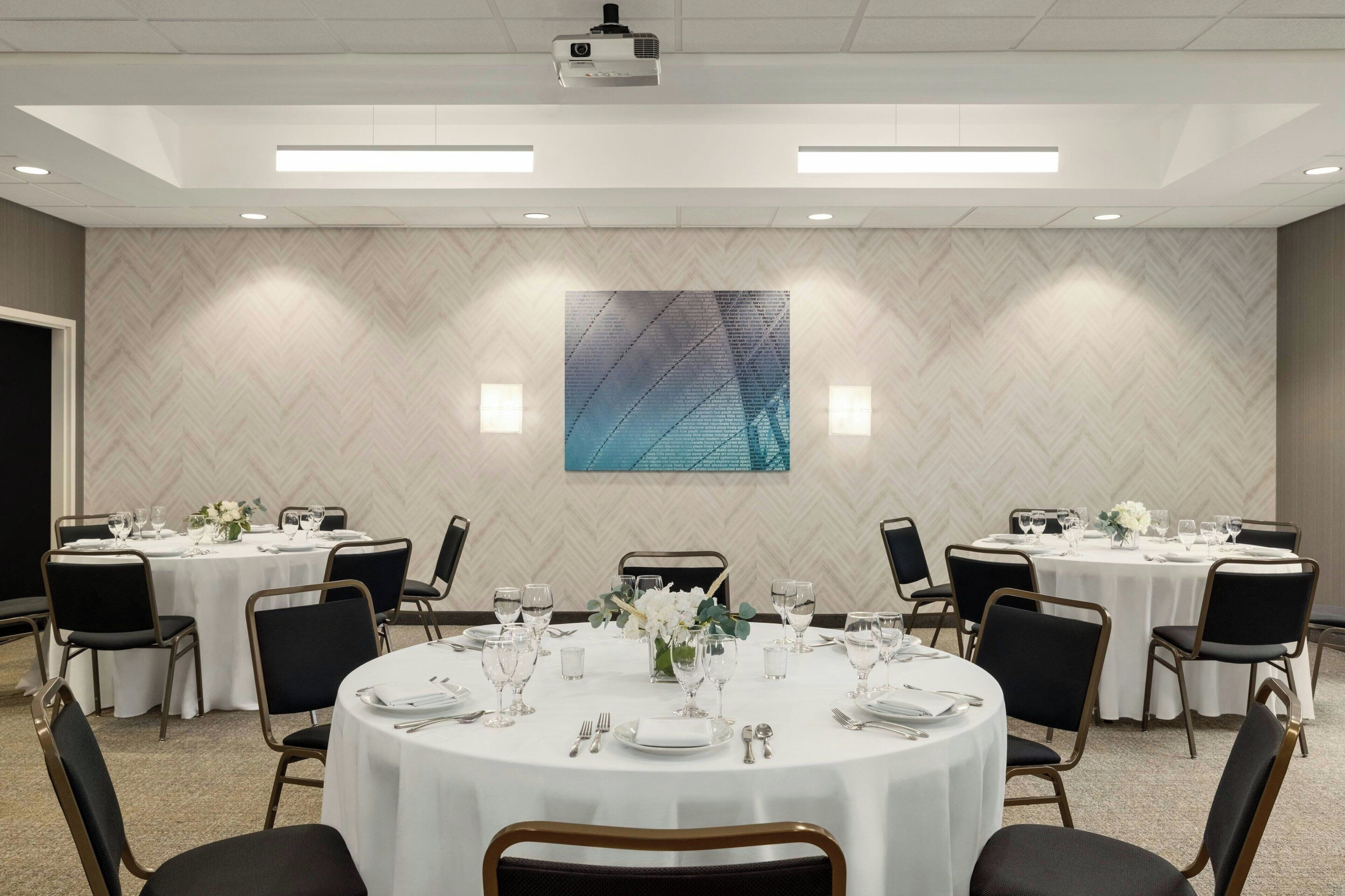 A conference room with round tables, black chairs, white tablecloths, and a large abstract painting on the wall.