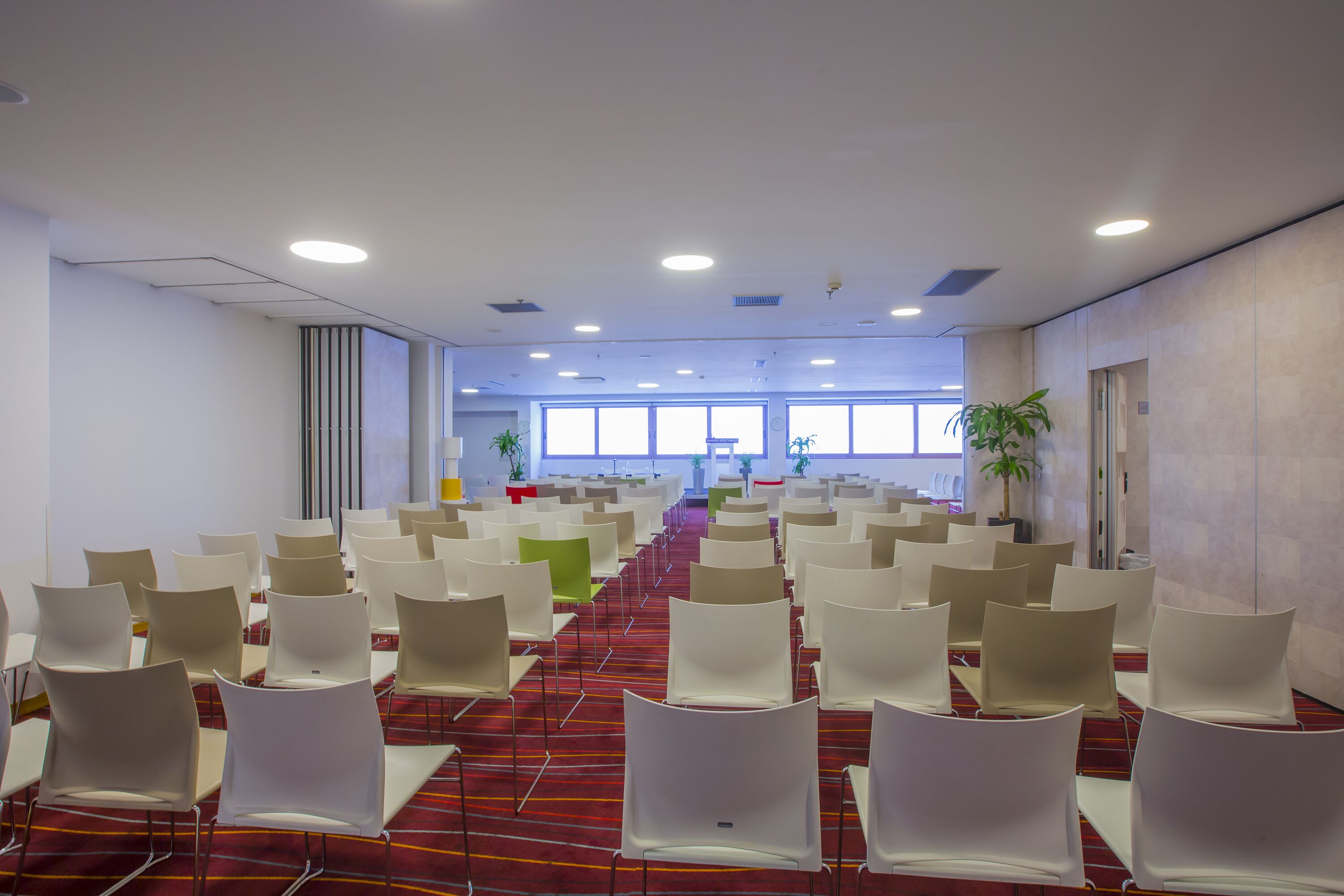 A conference room with rows of white chairs, a red carpet, and large windows allowing natural light.