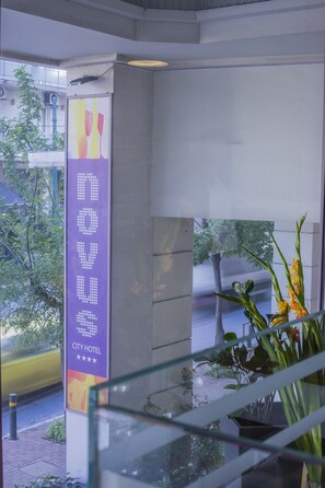 A modern hotel interior with a glass railing, a sign for the City Hotel, and a view of the street outside. - Novus City Hotel (Athens)