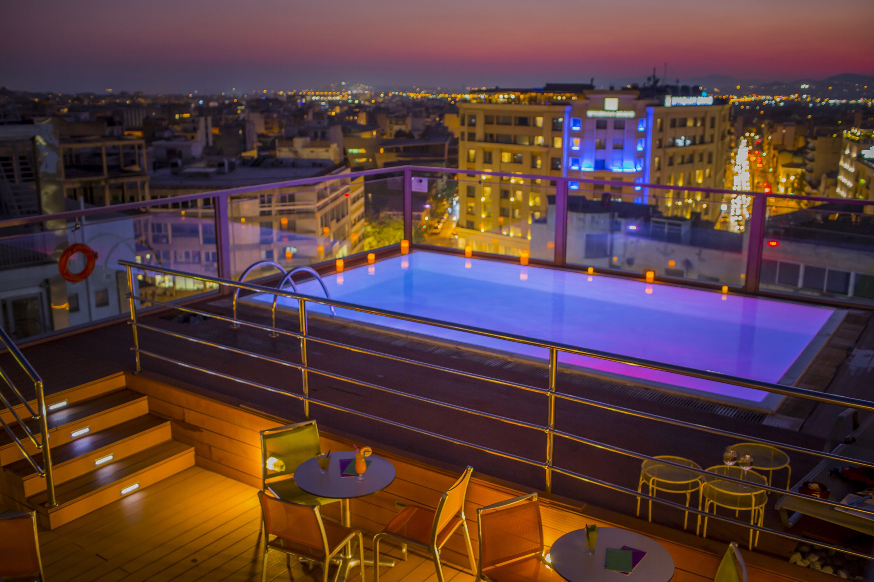 Una piscina en la azotea con barandillas de cristal, rodeada por una terraza de madera y sillas de metal.