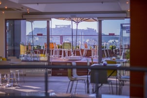 Ένας εξωτερικός χώρος για φαγητό, με τραπέζια και καρέκλες, μεγάλες ομπρέλες και θέα σε ένα αστικό τοπίο με κτίρια και έναν λόφο στο βάθος.