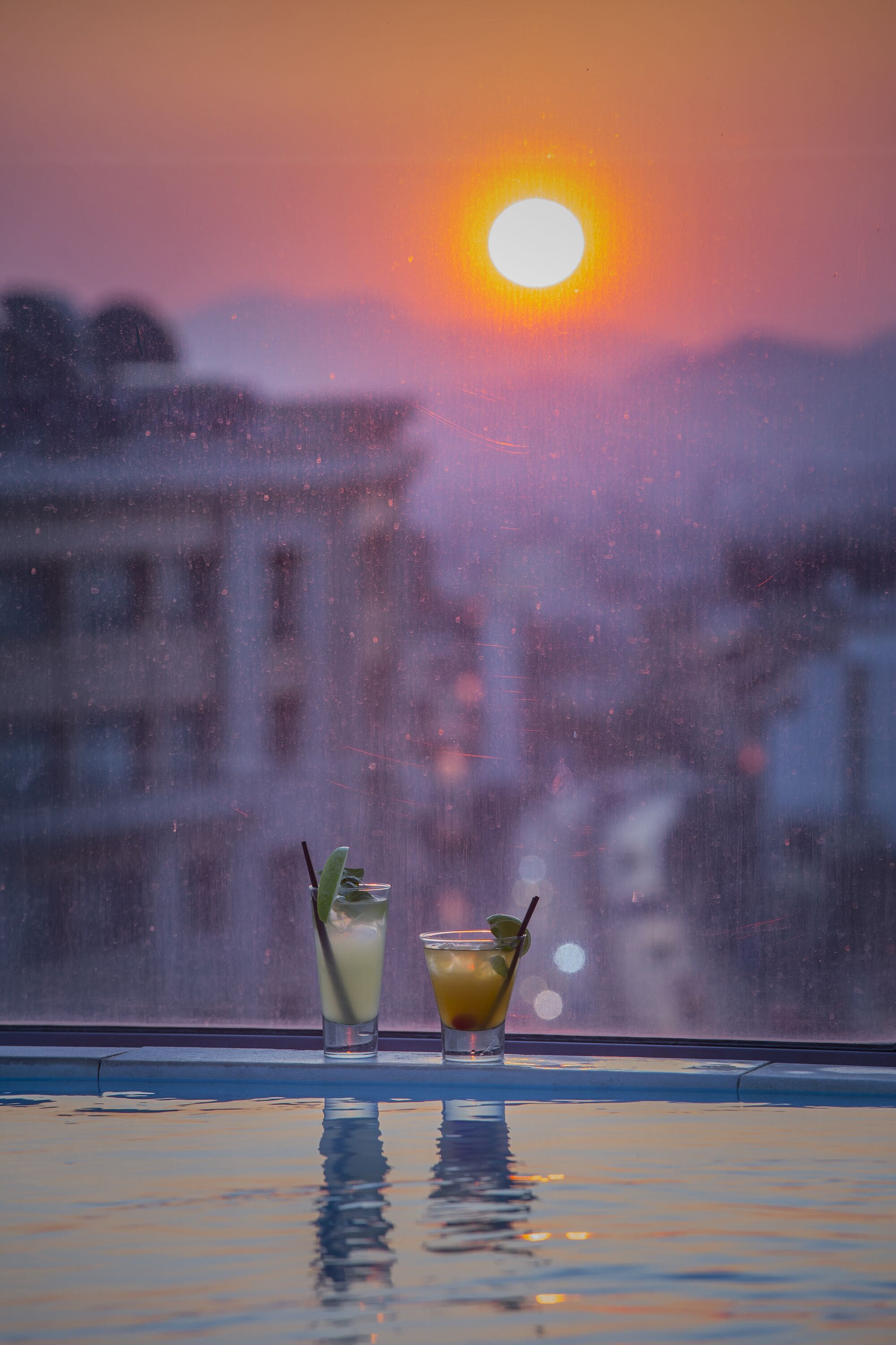 Two glasses of beverages on a reflective surface with a sunset view through a window.