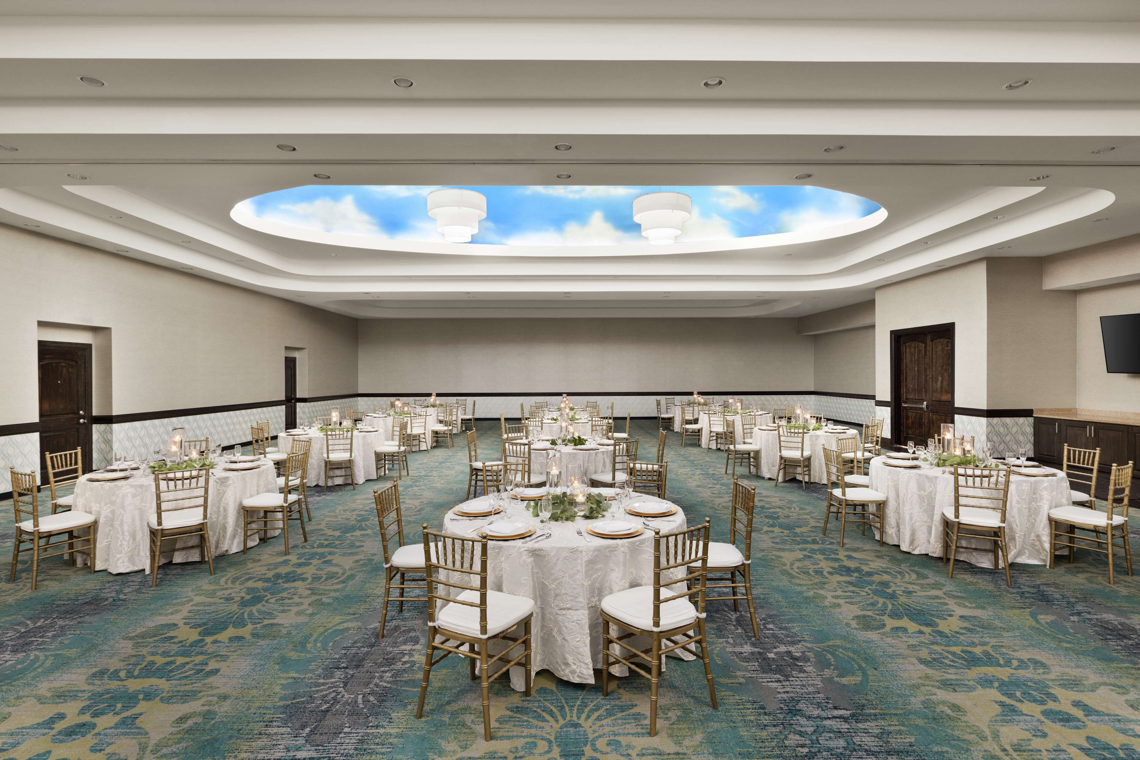 A large, well-lit banquet hall with multiple round tables set for dining, featuring white tablecloths and gold chairs.