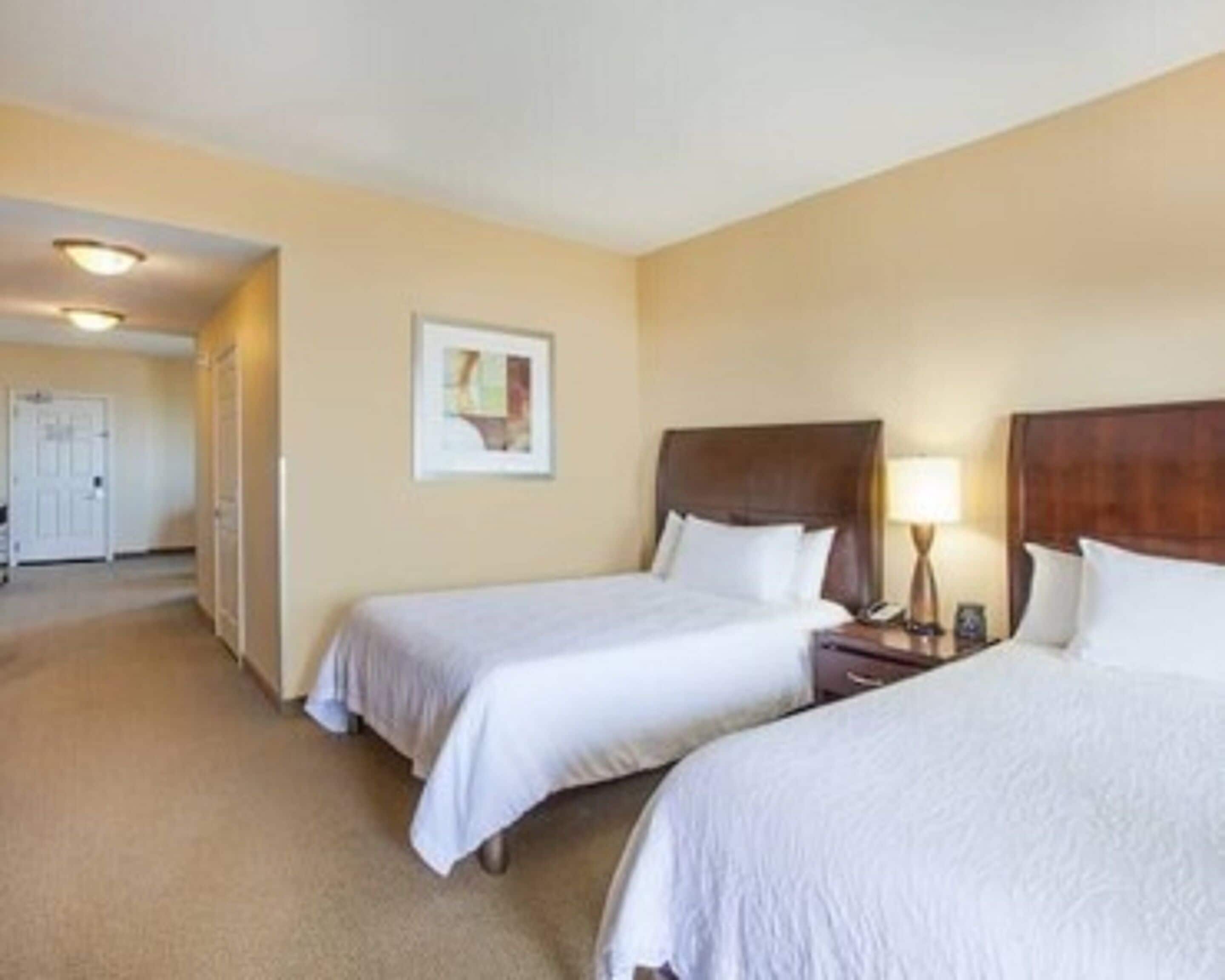 A hotel room with two single beds, a nightstand with a lamp, a framed picture on the wall, and a door leading to another room.