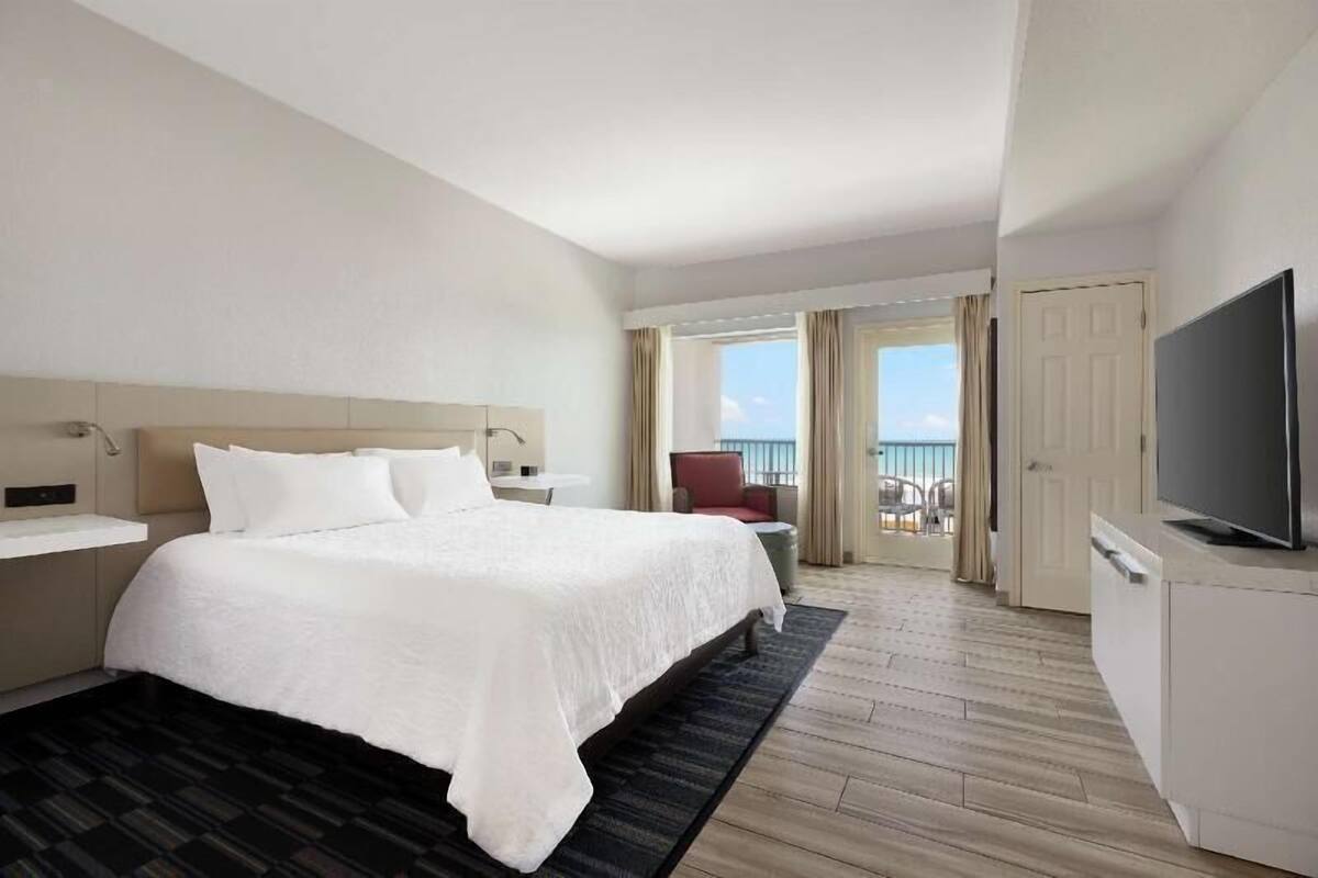 A hotel room with a large bed, a TV, a balcony with a view, and a door.