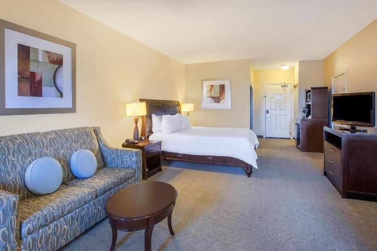 A hotel room with a bed, a sofa, a coffee table, a TV, and artwork on the walls.