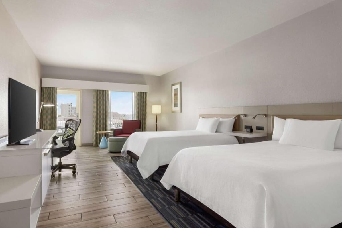 A hotel room with two beds, a desk with a chair, a TV, and a view of the city.