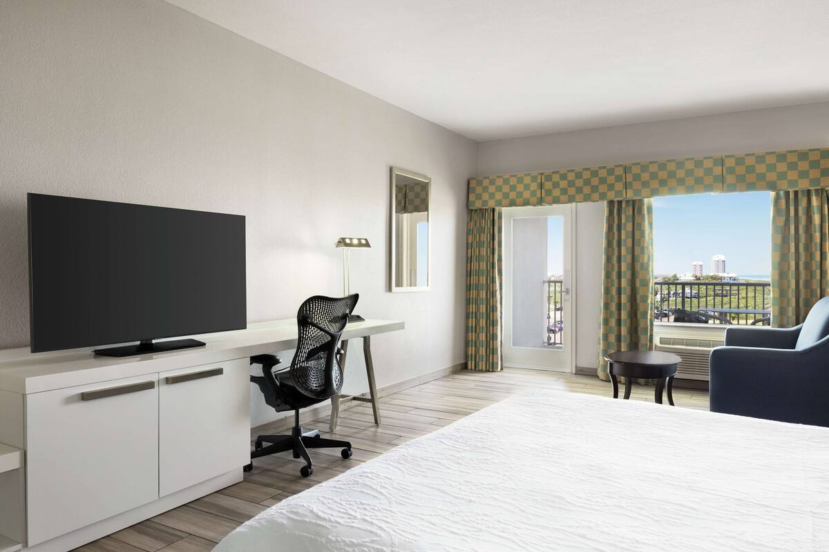 A hotel room with a bed, a desk with a television, a chair, a small table, and a balcony with curtains.