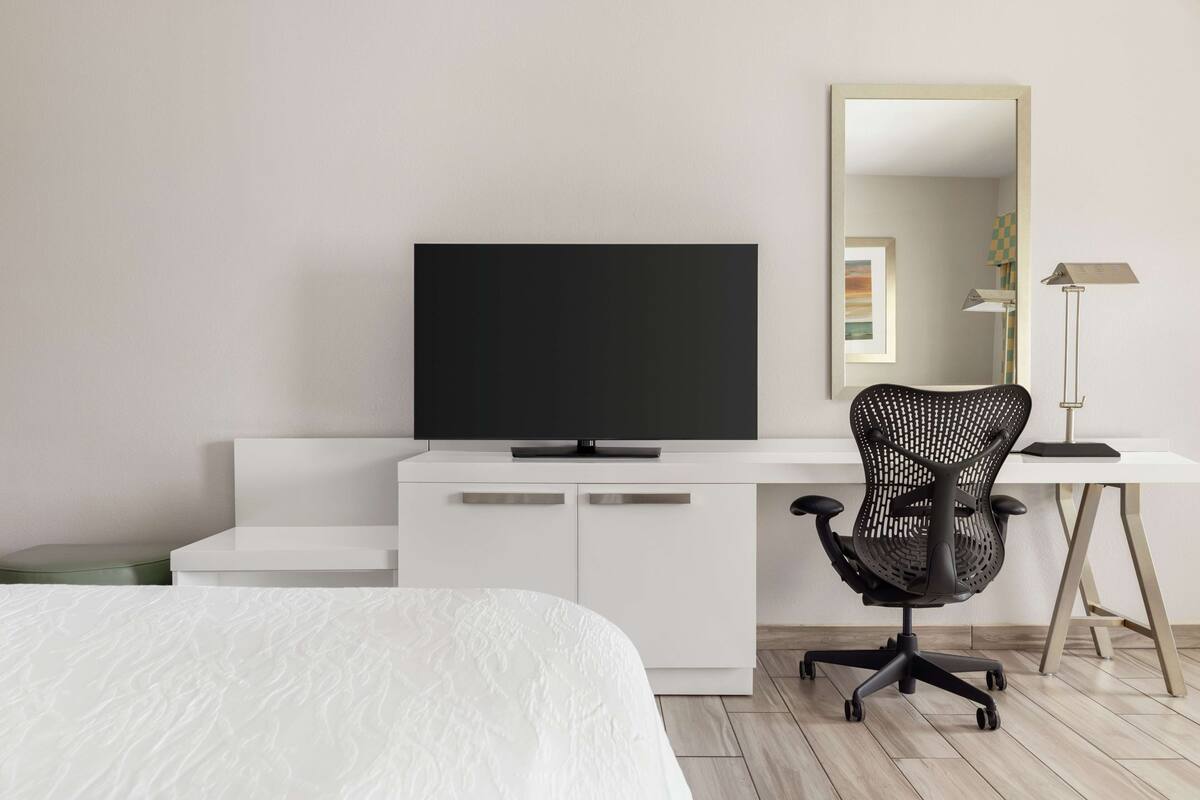 A modern bedroom with a bed, a white desk with a flat-screen TV, a black office chair, and a standing lamp.