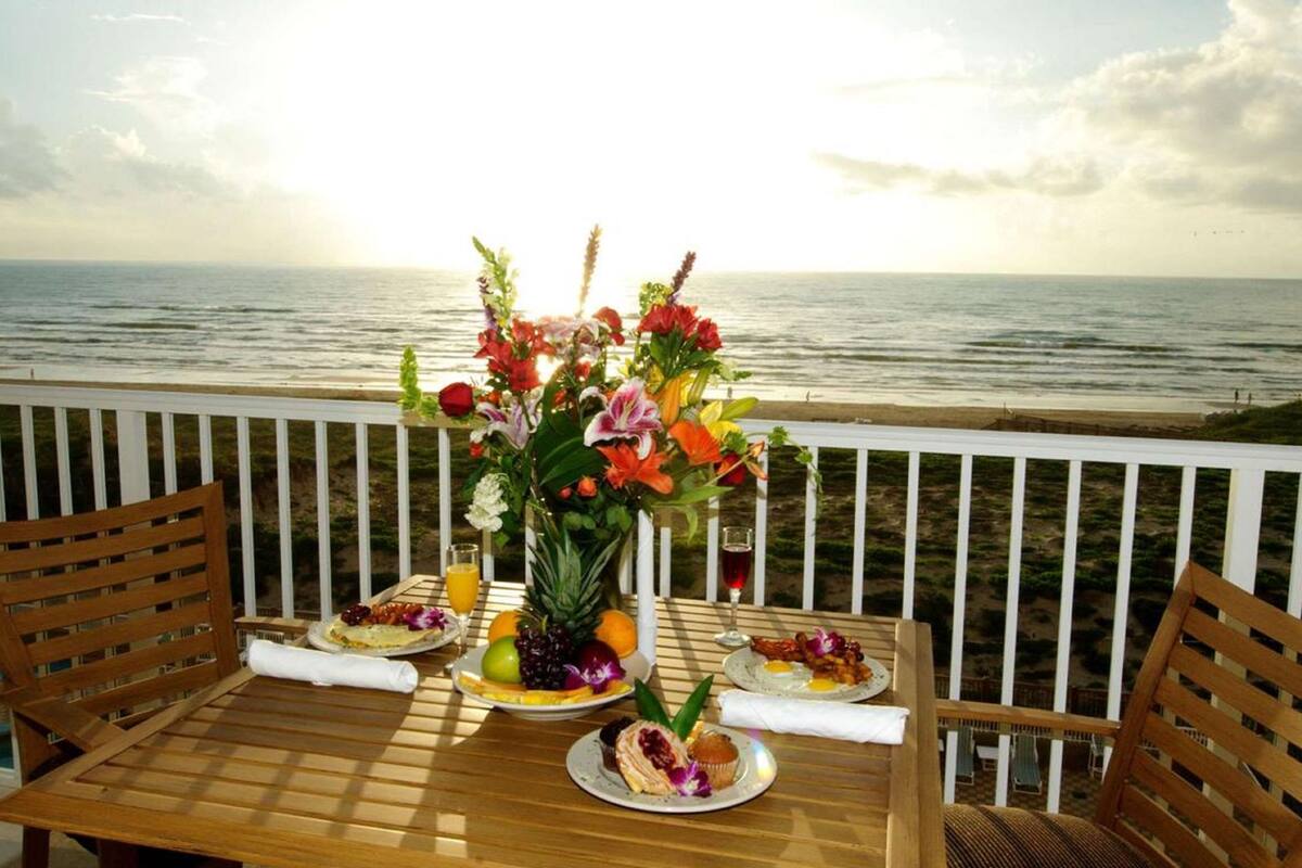 A wooden table with a floral centerpiece, plates of food, and drinks on a balcony overlooking the ocean.