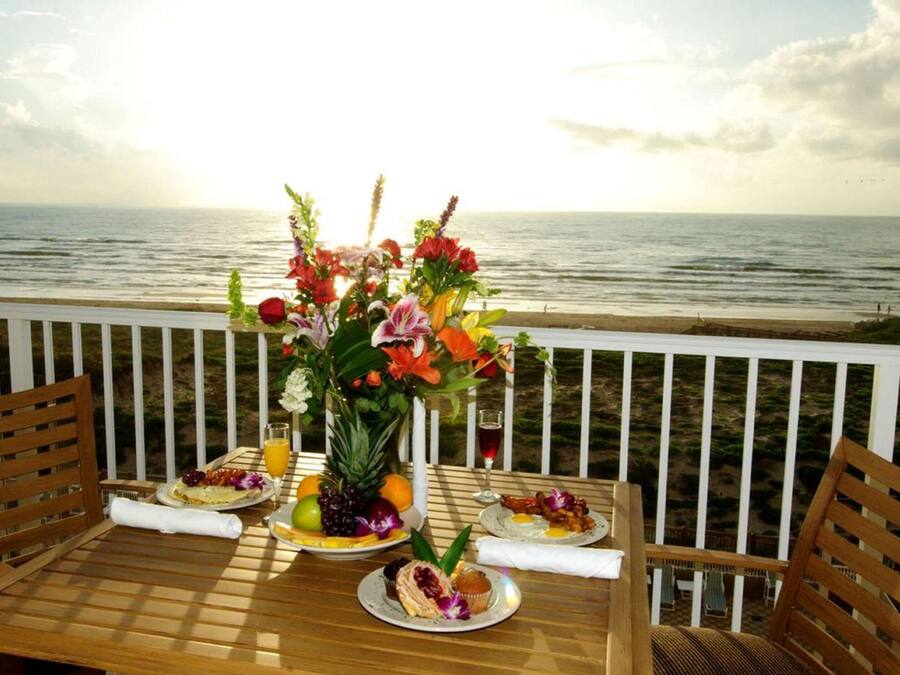 A wooden table with a floral centerpiece, plates of food, and drinks on a balcony overlooking the ocean.