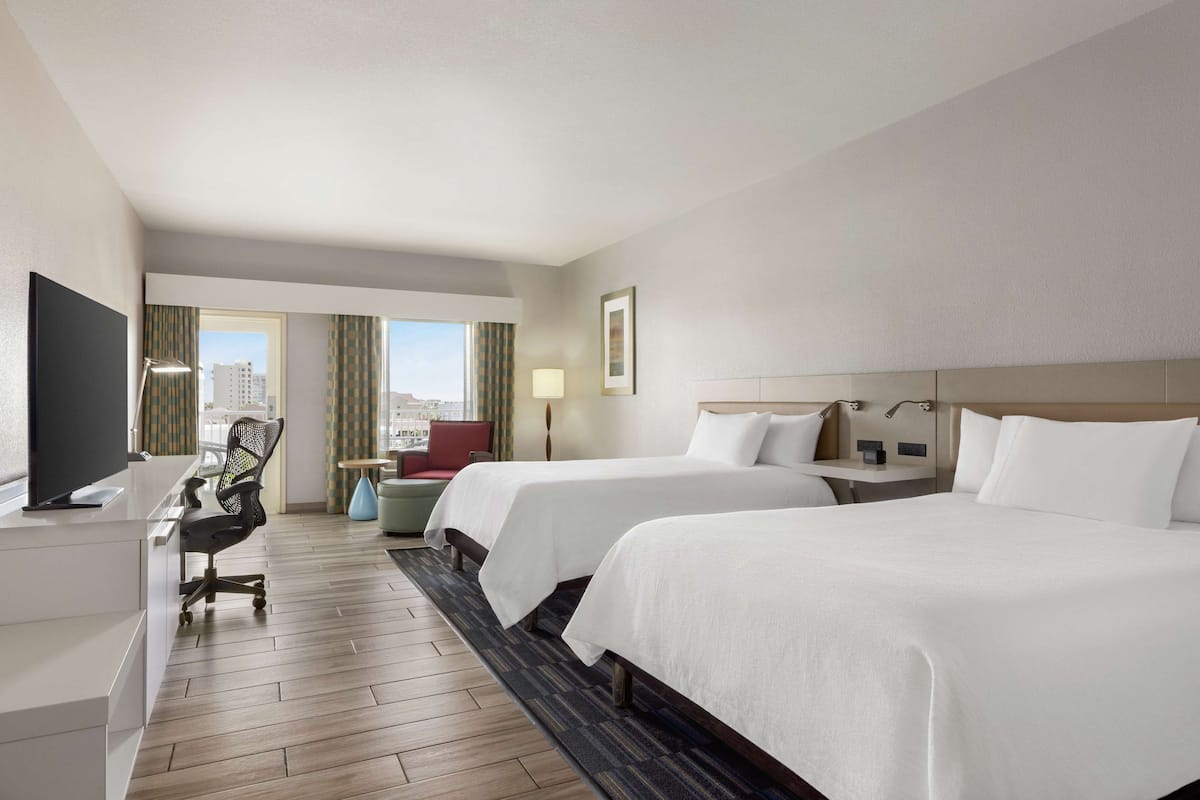 A hotel room with two beds, a desk with a chair, a TV, and a view of the city.